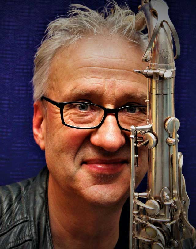 Das Foto zeigt das Portrait des Saxofonisten Anselm Simon. Er hält ein Saxofon neben seinem Gesicht, so dass ein kleiner Teil verdeckt wird. Er blickt lächelnd in die Kamera.
