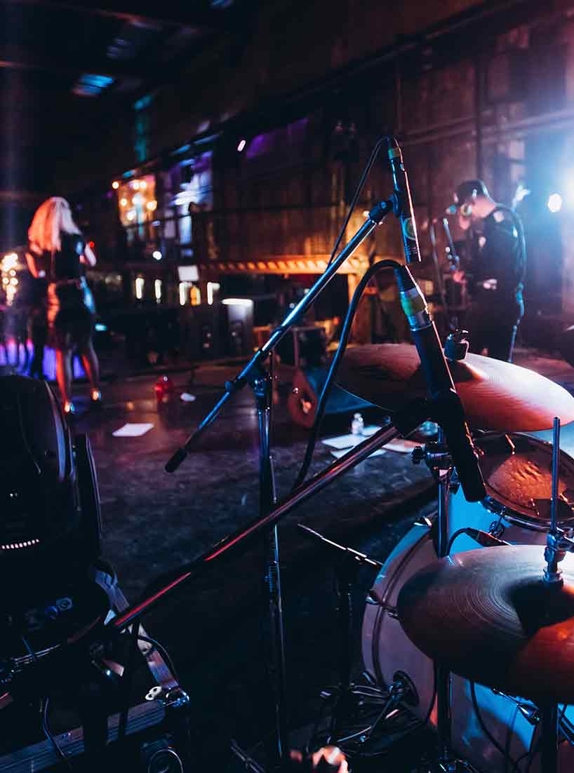 Auf eine Bühne stehen 2 Musiker, ein Gitarrist und eine Sängerin, beide sind von hinten zu sehen. Im Vordergrund befindet sich der Ausschnitt eines Schlagzeugs. Die Lichtstimmung ist schummrig mit bunten Lichtspots.