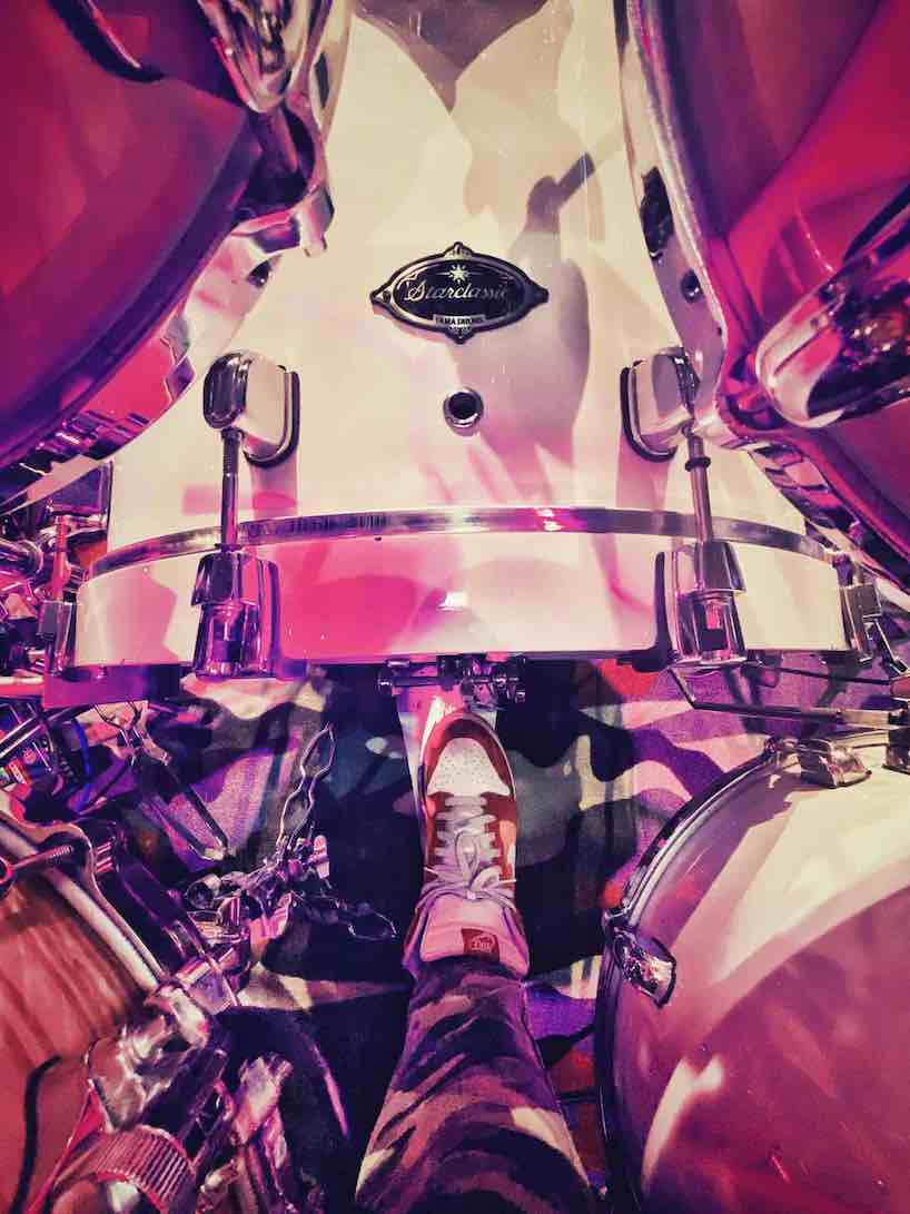 Eine ungewöhnliche Perspektive eines Schlagzeugs und Schlagzeugers aus der Vogelperspektive. Man sieht nur einen Ausschnitt, von dem Musiker nur einen Fuss. Insgesamt ist die Lichtstimmung sehr poppig und pink.