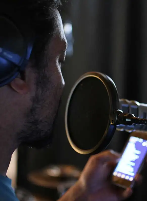 Man sieht das Portrait eines Musikers mit Kopfhörern und Mikrofon während einer Studioaufnahme von der Seite. Er hält ein Smartphone in der Hand, sein Blick geht Richtung Smartphone auf dem sehr unscharf ein Gesangstexte angedeutet wird.